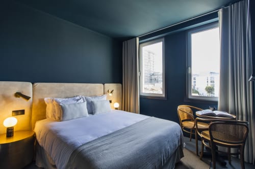 Quarto moderno de hotel no Porto com paredes azul escuro, cama queen com iluminação aconchegante, grandes janelas com vista para a cidade e espaço de trabalho