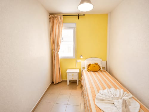 Dormitorio amarillo brillante con cama individual, ropa de cama naranja a rayas, mesita blanca y ventana en Apartamentos El Calón Playa