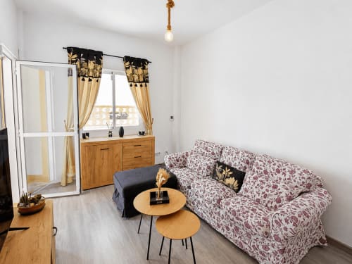 Luminoso salón con sofá floral, mesas de madera y elegantes cortinas doradas en Apartamentos El Calón Playa,