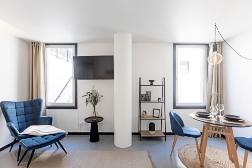 Apartamento estúdio moderno no Porto com cadeiras acolchoadas azuis, mesa de jantar, coluna branca, TV de tela plana e luz natural
