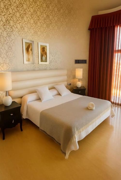 Elegante suite con cama king en Hotel Villa de Cacabelos: ropa de cama crema, papel tapiz decorativo, cortinas rojas y piso de madera