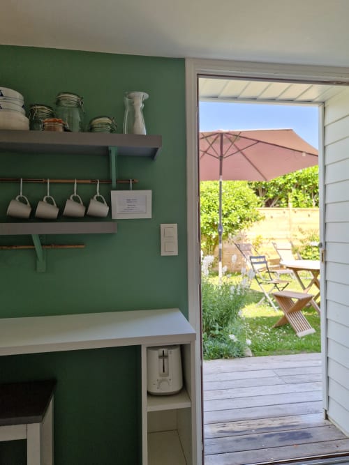 Kitchenette moderne aux murs verts, étagères ouvertes de rangement, vue sur la terrasse jardin des Chambres du Keriolet