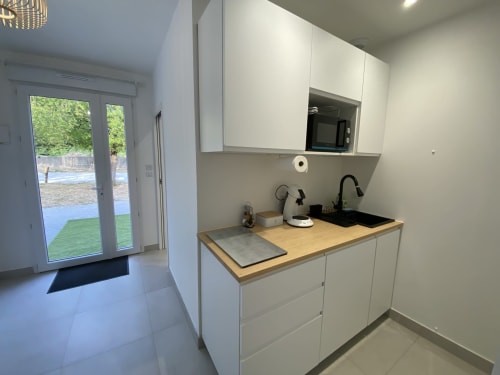 Modern kitchenette in La Maison Blanche apartment with white cabinets, wooden countertop, and garden view through patio doors