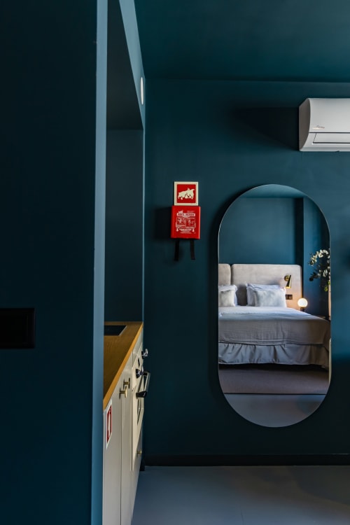 Quarto em tons azul-esverdeado com espelho oval refletindo a cama, extintor vermelho na parede, ar condicionado, estúdio moderno de hotel no Porto