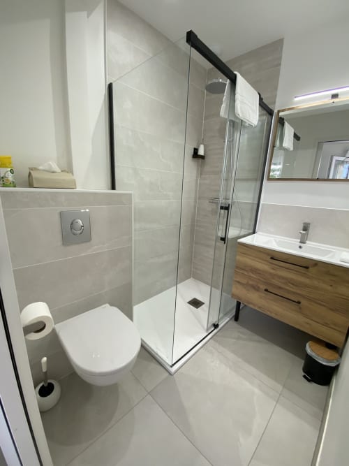 Salle de bain moderne du studio La Maison Blanche avec douche vitrée, toilettes blanches, meuble vasque en bois et radiateur-sèche-serviettes