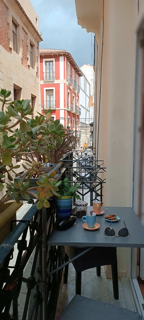 Vista del balcón de la Pensión Alicante San Nicolás con edificio de rayas rojas y plantas en macetas sobre la mesa