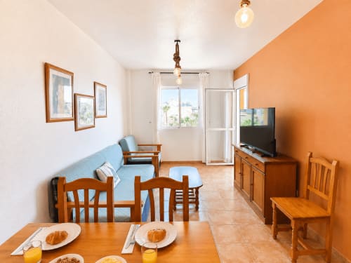 Open-plan living area in Apartamentos El Calón Playa with dining table, blue sofa, TV, and sea-view balcony in Cuevas del