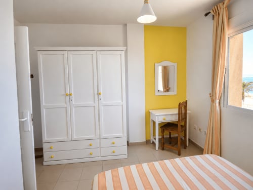 Luminoso dormitorio con armario blanco, cama de rayas, pared amarilla y vistas al mar en Apartamentos El Calón Playa