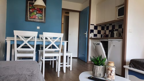 Modern apartment dining area with white chairs, blue walls, kitchen, and sea view at Ô CAP COZ in La Fôret-Fouesnant