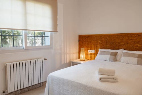 Dormitorio luminoso de la cabaña Las Golondrinas con cabecero de madera, cama blanca, ventana con vista al jardín y radiador de calefacción en Cap