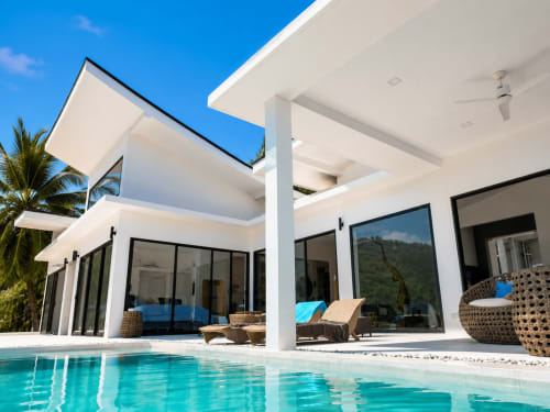 Modern white villa in Ko Samui with turquoise pool, lounge chairs, and tropical palm trees under clear blue sky