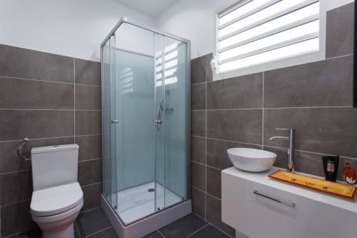 Salle de bains moderne du gîte Kaz A Bibi à Sainte-Rose avec douche vitrée, lavabo blanc et murs carrelés foncés