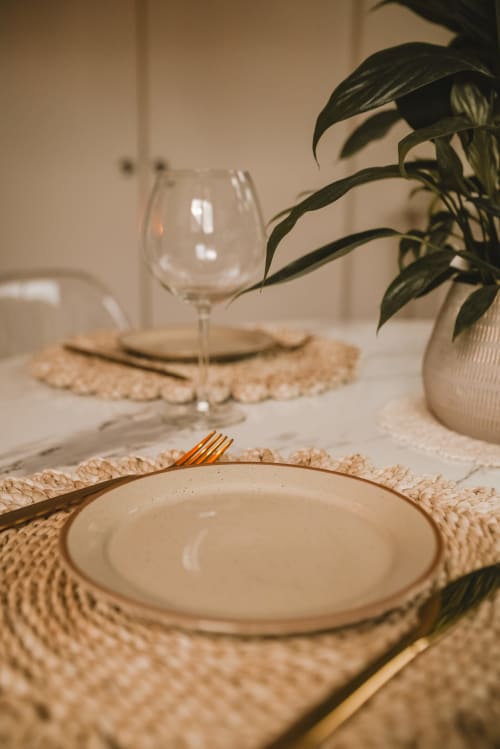 Mise en place élégante au cottage ART DE VOYAGER à Étretat : assiette crème, fourchette dorée, verre à vin et plante en pot sur tissage