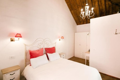 Dormitorio luminoso en Casa 1820 de Santa Cruz de la Palma con cama blanca, almohadas rojas, cabecero ornamentado y techo de madera