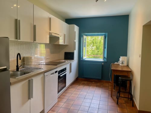 Cuisine de charme avec carrelage terracotta, meubles crème, mur accent bleu-vert et vue sur le jardin au Château de Vaux