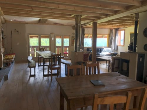 Open-plan living area in mountain chalet with wooden dining tables, fireplace, and large windows overlooking Alps