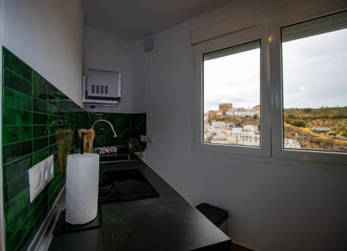 Moderne Küche mit grünen Fliesen, schwarzen Arbeitsplatten und großem Fenster mit Blick auf das weiß getünchte Dorf Setenil de las Bodegas