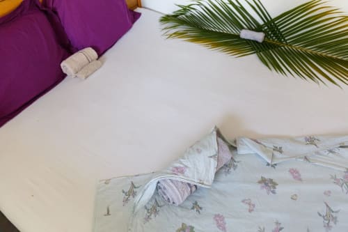 Lit cottage Kaz A Bibi avec coussins fleuris, draps blancs, tissu pourpre, frondes de palmier et serviettes roulées