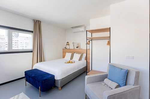 Quarto de estúdio moderno em hotel no Porto com roupas de cama brancas, banco azul, poltrona, prateleiras de madeira e vista para a cidade