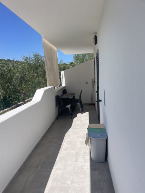 Patio stretto con muri bianchi, contenitori per il riciclaggio, tavolo e sedie neri, ulivi visibili, vista del giardino di Villa Angela