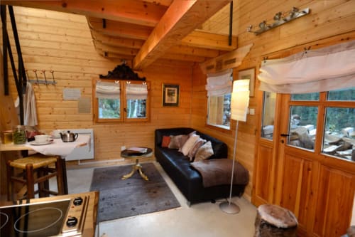 Intérieur cosy du chalet en bois à l'Auberge Nemoz : canapé noir, kitchenette, et vue montagne par les grandes fenêtres