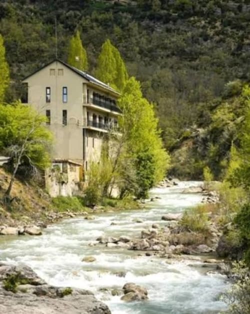 Edificio de apartamentos beige junto a un arroyo de montaña rodeado de altos cipreses en los Pirineos de Montgat