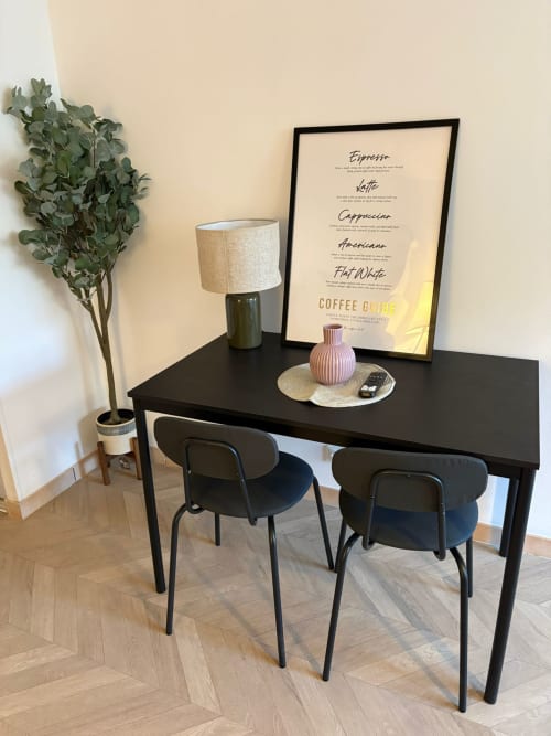 Bureau noir moderne avec deux chaises, lampadaire, affiche guide café et plante en pot dans l'appartement Cosy Luxe du Paradis Des