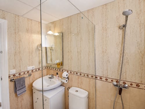 Baño moderno con paredes alicatadas en beige, lavabo blanco, inodoro, bidé y ducha con mampara de cristal en Apartamentos El Calón Playa,