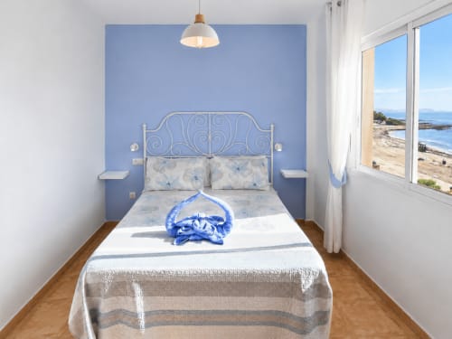 Bright beachfront apartment bedroom with blue accent wall, ornate metal bed, sea view through terrace doors, Cuevas del