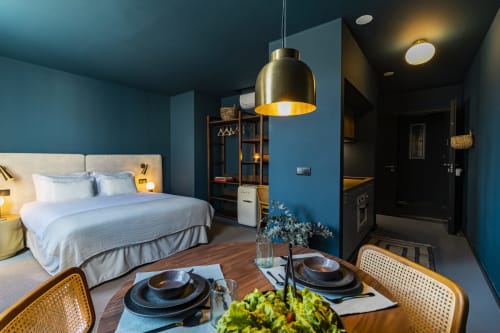 Estúdio moderno no Porto com paredes azuis, luminária pendente de latão, área de refeições, kitchenette e cama com lençol branco.