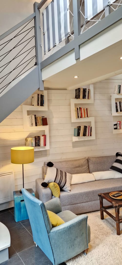 Salon moderne avec escalier gris, étagères intégrées blanches, canapé beige, fauteuil bleu et lampe jaune aux Chambres du