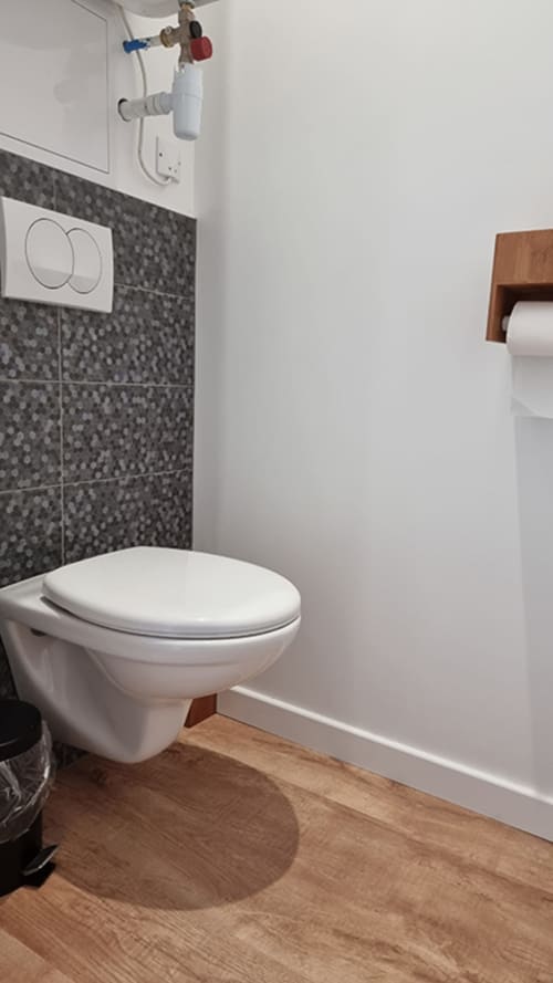 Modern bathroom with wall-mounted toilet, dark tile accent wall, wooden flooring, and white walls at Ô LOCH apartment, La