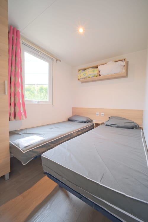 Chambre moderne de mobilhome avec deux lits, rideaux roses et mobilier en bois à Annick Camping, Vias Plage