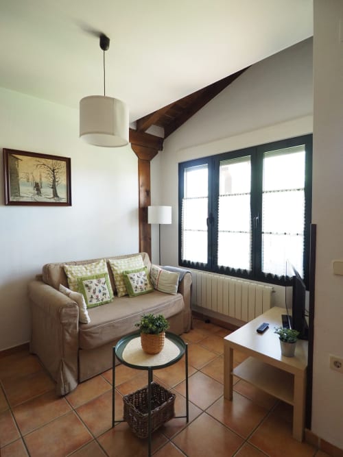 Acogedora sala de estar en apartamento rural español con sofá beige, mesita redonda y vistas al jardín por ventanas de marco oscuro