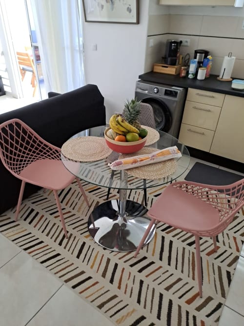 Salle à manger contemporaine de l'appartement Kay Betty en Martinique avec chaises roses, table ronde en verre, coupe de fruits et tapis rayé