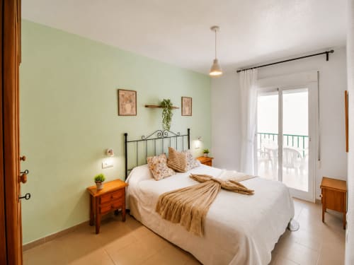 Dormitorio luminoso con pared verde pálido, ropa de cama blanca, cama de hierro forjado, mesita de noche de madera y acceso a balcón