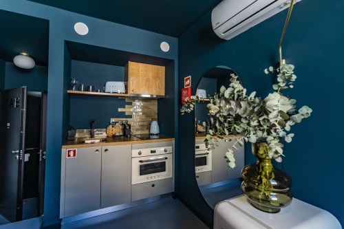 Kitchenette moderna e compacta em hotel do Porto com paredes azul-petróleo, eletrodomésticos brancos e arranjo floral fresco.