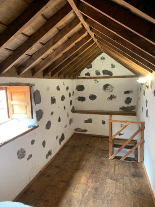 Dormitorio rústico en buhardilla con vigas de madera vista, paredes blancas con manchas de moho, suelo de madera y pequeña ventana en Casa