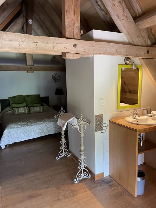 Chambre de gîte avec poutres apparentes, literie verte et salle de bain attenante avec miroir à cadre jaune aux Pratges, Figeac