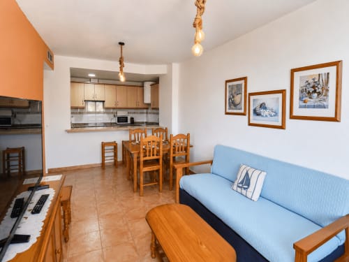 Open-plan apartment living area with wooden furniture, blue sofa, dining table, kitchen, and sea view artwork at Apartamentos