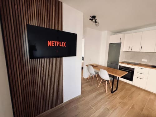 Cocina y comedor de apartamento moderno con Netflix en TV montada en pared, mesa de madera, sillas blancas y equipada