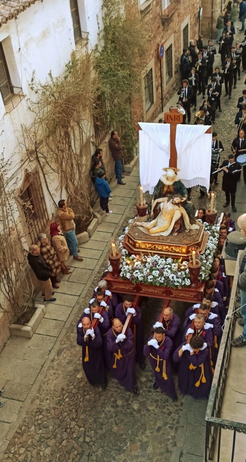 Procesión religiosa en la histórica calle de Cáceres cerca del Mirador Medieval con santuario ornamentado y participantes enrojecidos