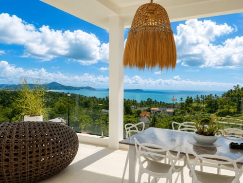 Tropical villa terrace overlooking Ko Samui's turquoise sea with white dining furniture and woven pendant light