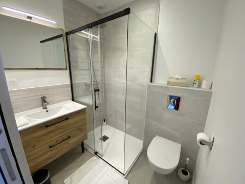 Salle de bain moderne de l'appartement La Maison Blanche à Bouguenais. Douche vitrée, meuble vasque en bois, toilettes blanches, murs carrelés.