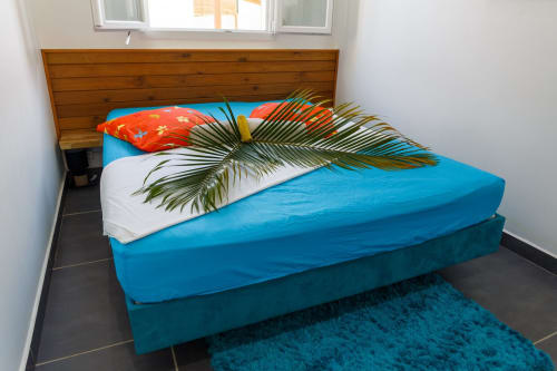 Chambre lumineuse du gîte Kaz A Bibi avec tête de lit en bois, literie turquoise, feuilles de palmier et carrelage foncé
