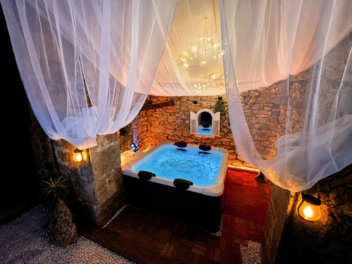 Luxueux bain à remous extérieur aux eaux bleues sous des voilages blancs aériens, murs en pierre, éclairage chaleureux au Domaine Valenquercy