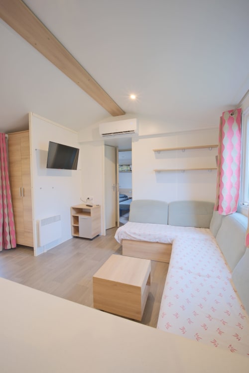 Intérieur moderne de mobil-home avec lits jumeaux, télévision, plafond à poutres en bois et rideaux roses au camping Annick, Vias Plage