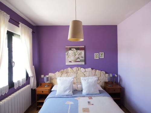 Dormitorio acogedor con paredes moradas, cabecero de piedra, almohadas blancas y lámpara colgante en apartamento rural de Sebúlcor