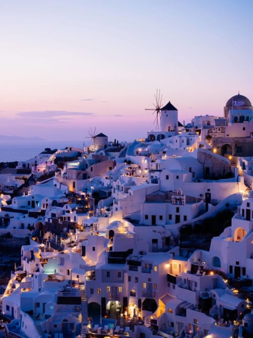Pueblo de Santorini al atardecer con casas encaladas, molinos de viento e iglesia de cúpula azul iluminada al anochecer