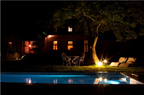 Le Petit Manoir de Kériolet illuminé la nuit, bâtiment en briques étincelant, piscine lumineuse, chaises longues et cour éclairée